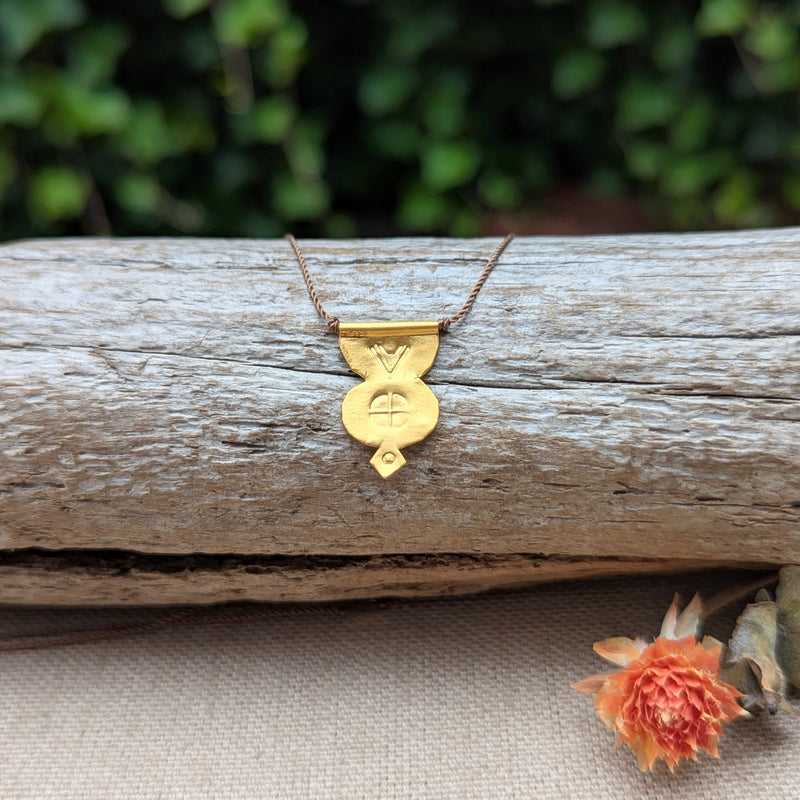 Gold necklace with a pendant and a brown cord on a wooden log with a blurred green background