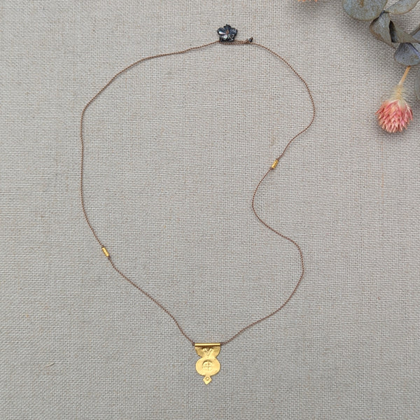Gold necklace with a pendant with a textured design and a brown cord with a black flower bead clasp on a textured beige surface