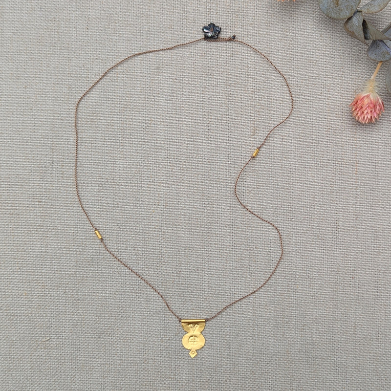 Gold necklace with a pendant with a textured design and a brown cord with a black flower bead clasp on a textured beige surface
