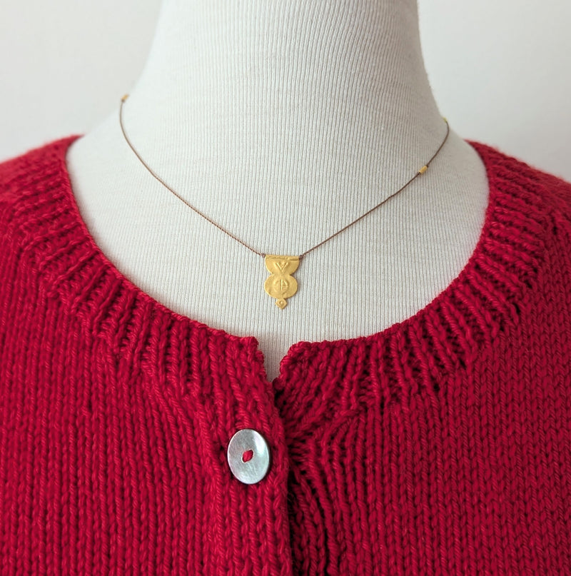 Gold pendent necklace with a textured design on a brown cord on a mannequin with a red knitted cardigan