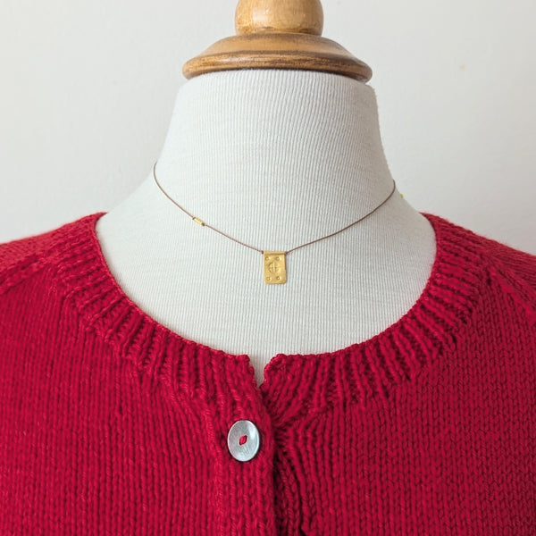 Red knitted cardigan on a mannequin with a necklace with a gold rectangular pendant with a textured design on a tan cord with a white background. 