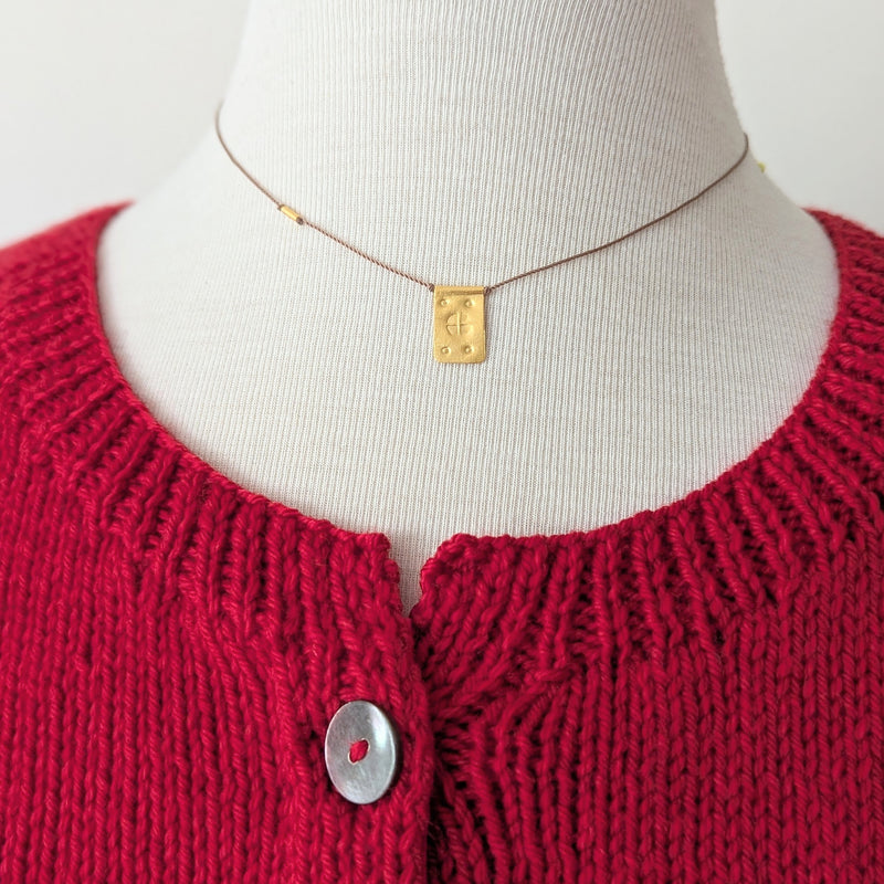 Red knitted cardigan on a mannequin with a gold necklace featuring a rectangular pendant with a textured design on a tan cord.