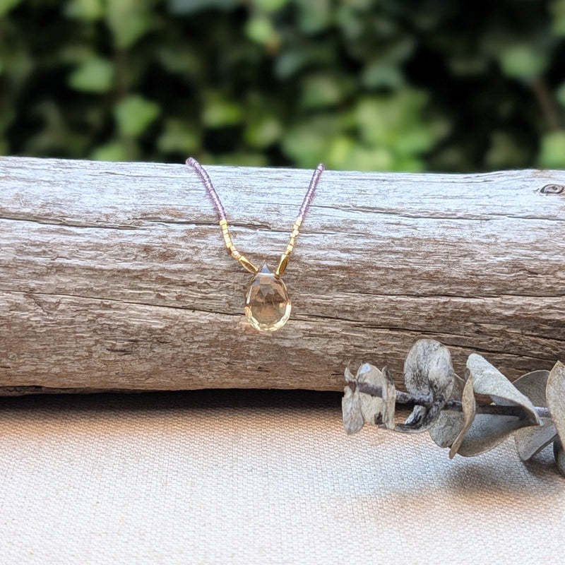 Liliac and gold beaded necklace with a quartz pendant on a wooden log with eucalyptus leaves