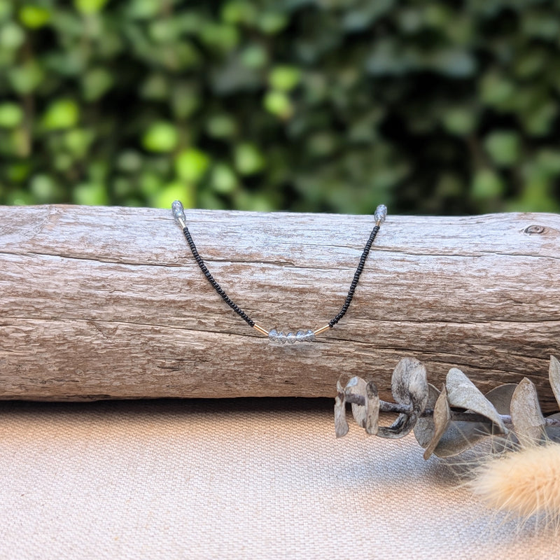 Necklace with small black, grey, and gold beads on a wooden log with a blurred green background