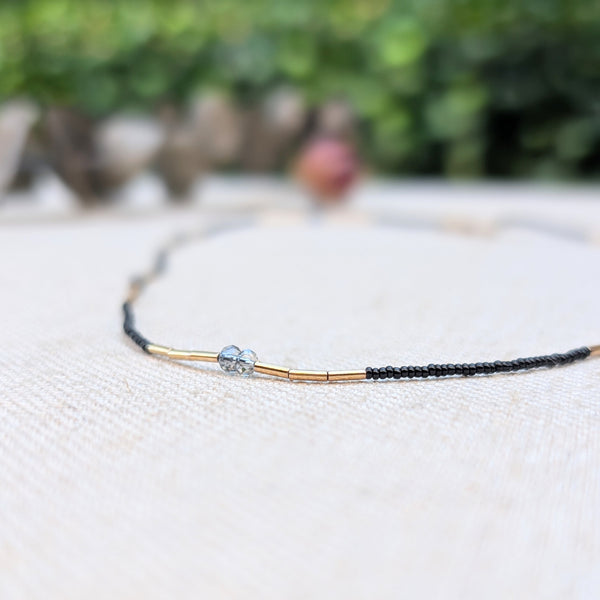 Delicate gold, grey, and black beaded necklace on a textured surface with a blurred natural background