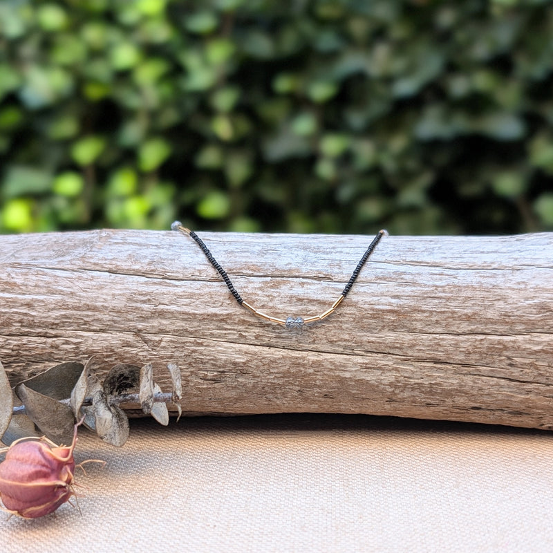 Necklace with small grey, gold, and black beads on a wooden log with a blurred green background