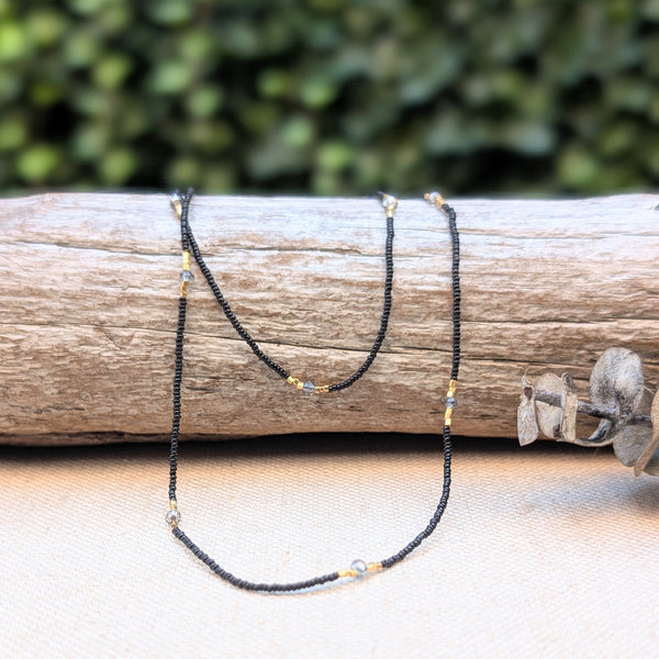 Black, grey, and gold beaded necklace on a wooden log with a blurred gree background.