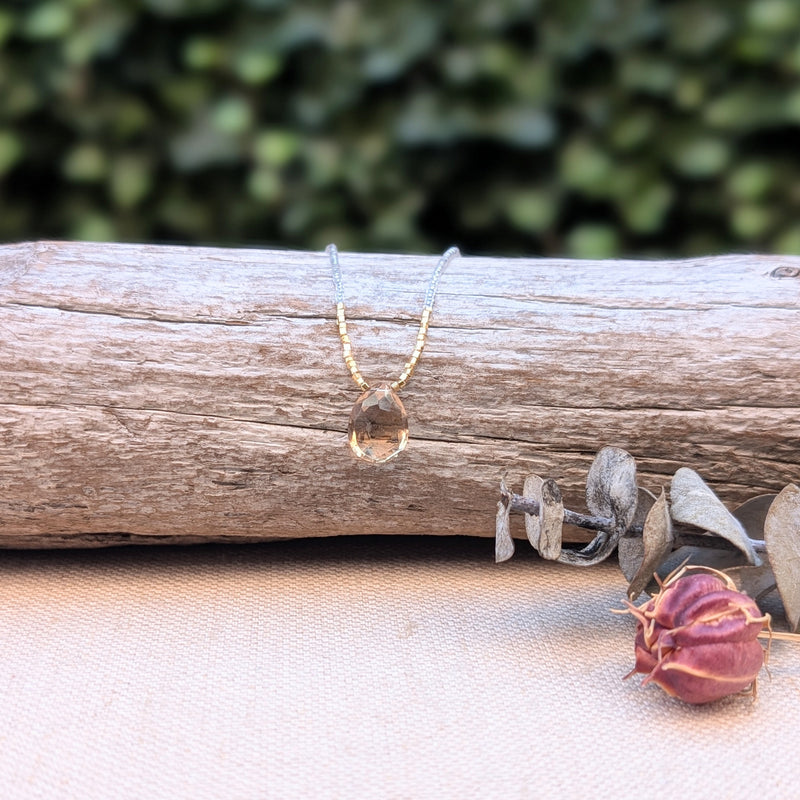 Clear and gold beaded necklace with a teardrop-shaped pendant on a wooden log with greenery in the background