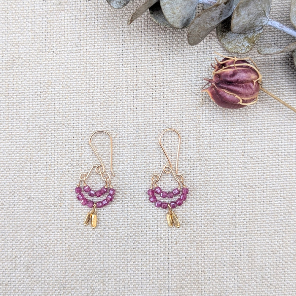 gold earrings with ruby beads hanging off in two rows on a white textured background with dried flowers on the side
