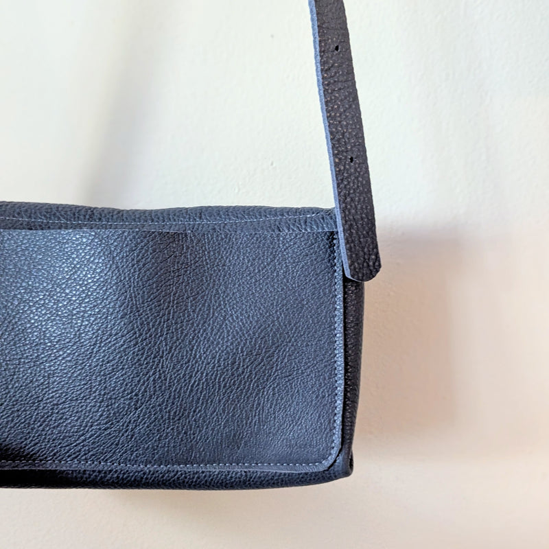 Back pocket of blue Stitch and Tickle mini messenger crossbody bag against a white wall.