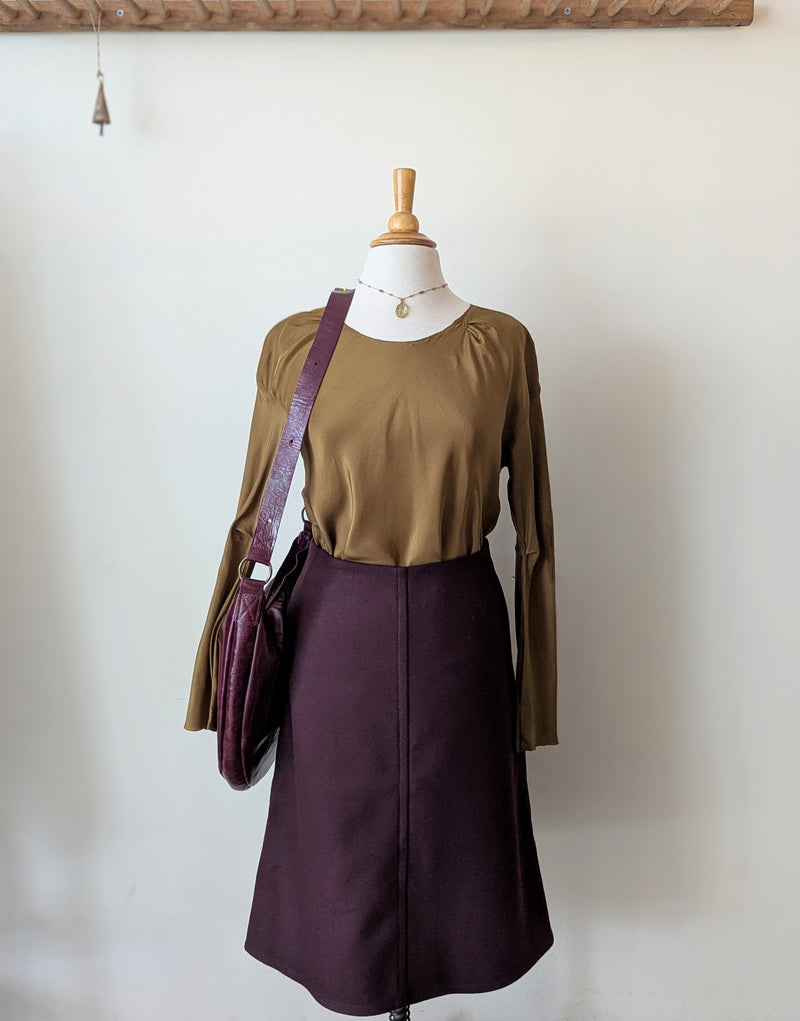 Mannequin wearing a green top and purple skirt with a Stitch and Tickle Luna Bag in Wine  against a white wall.