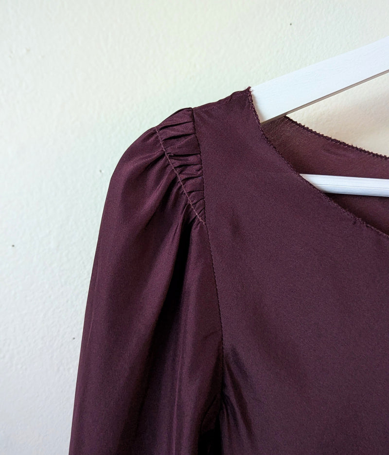 Close-up of a burgundy Susanne bommer blouse with ruched shoulder detail on a white hanger against a beige wall.
