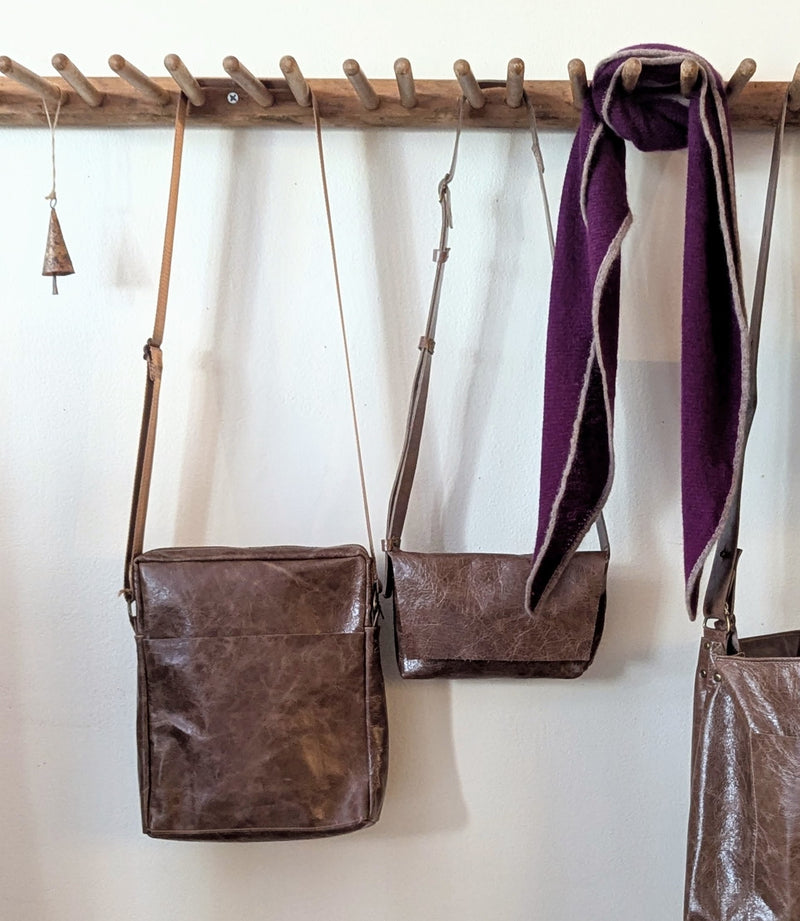 Brown Stitch & Tickle leather bags and a purple scarf hanging on a wooden rack against a white wall.