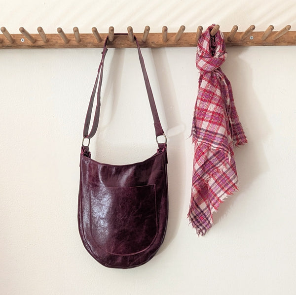 Stitch and Tickle Luna Bag in Wine and plaid scarf hanging on a wooden rack against a white wall.
