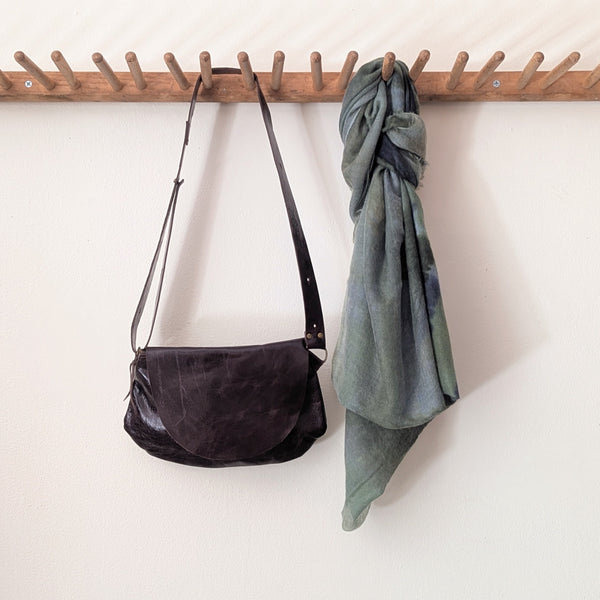 Stitch & Tickle Passito leather bag in Espresso and green scarf hanging on a wooden rack against a white wall.