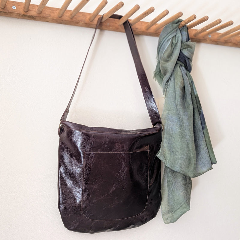Stitch and Tickle Passy Bag in Espresso  and green scarf hanging on a wooden rack against a white wall.