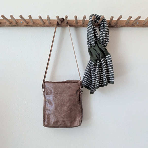Stitch & Tickle Bourvil Bag in Chai and a green and white checkered scarf hanging on a wooden rack against a white wall.