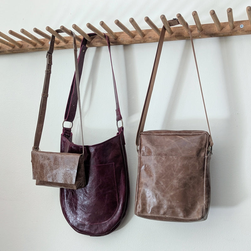 Three Stitch & Tickle leather bags hanging on a wooden rack against a white wall.