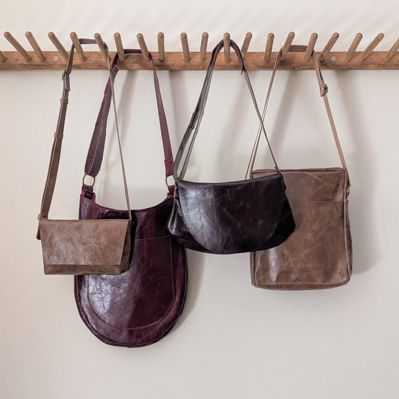 Four leather Stitch & Tickle handbags of varying sizes hanging on a wooden rack against a white wall.