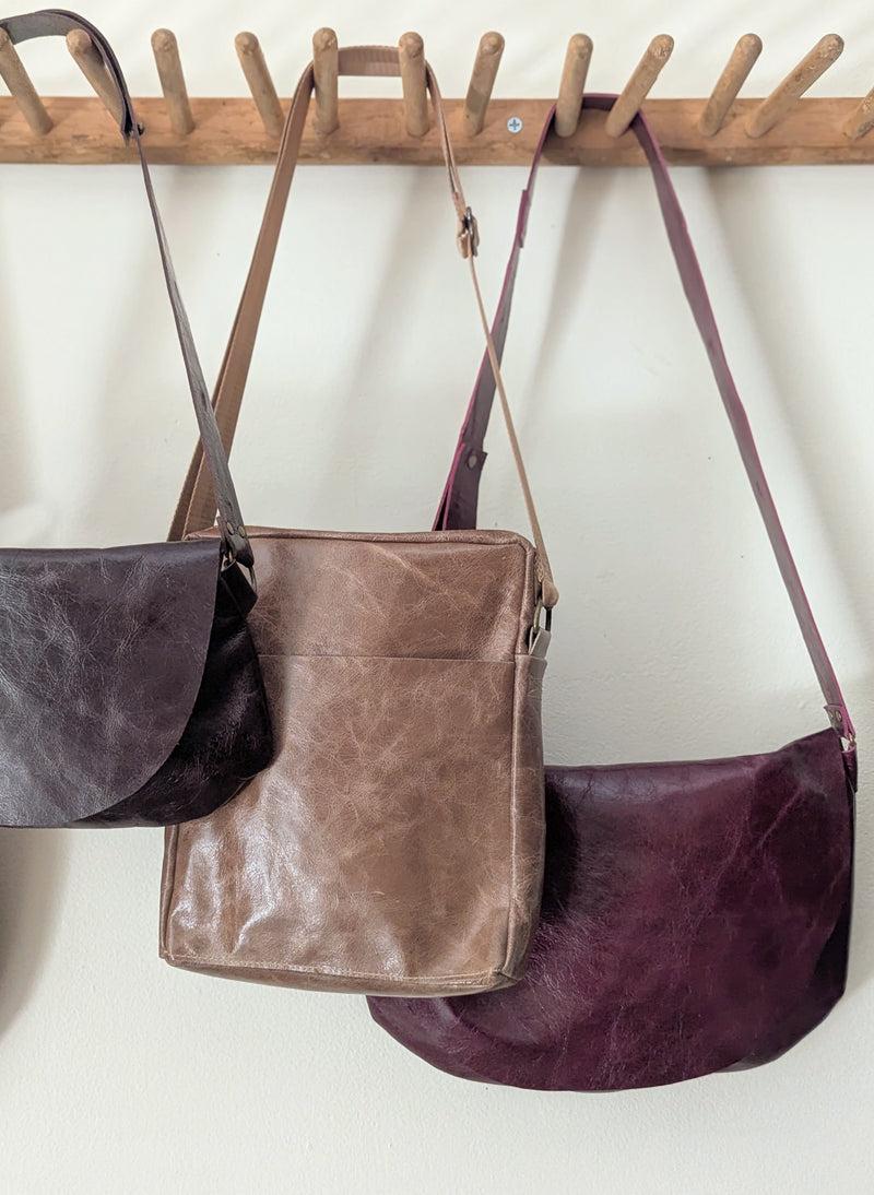 Three leather Stitch & Tickle bags hanging on a wooden rack against a white wall.