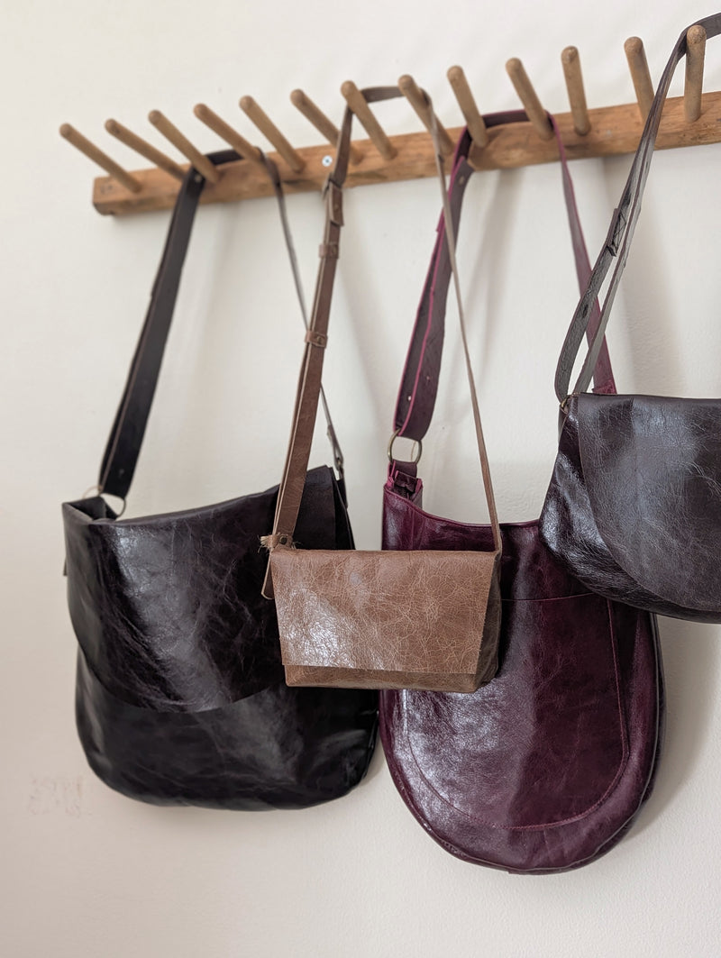Four leather Stitch & Tickle handbags hanging on a wooden rack against a white wall.
