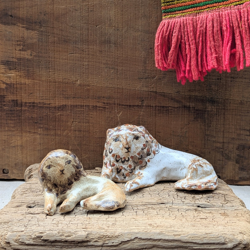 Two ceramic lion sculptures on a wooden surface with a textured wall and pink fringe in the background.