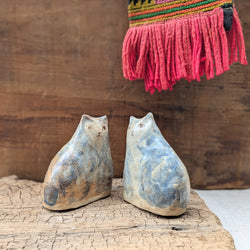 Two ceramic cat sculptures on a wooden surface with a colorful woven scarf in the background.