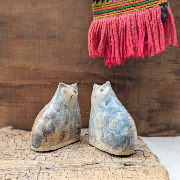 Two ceramic cat sculptures on a wooden surface with a colorful woven scarf in the background.