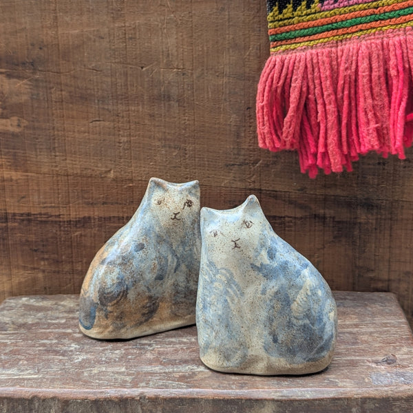 Two ceramic cat sculptures on a wooden surface with a colorful woven item in the background.