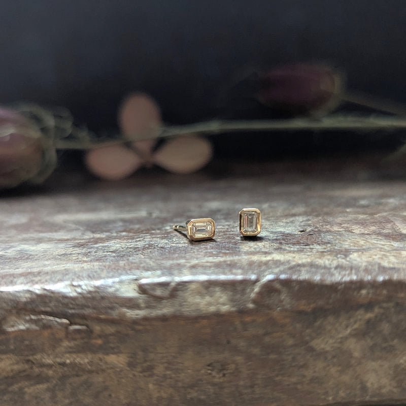Gold emerald-cut diamond stud earrings on a wooden surface with blurred flowers in the background