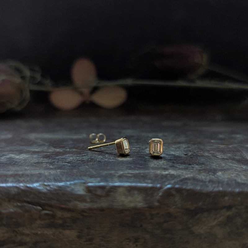 Gold stud diamond earrings on a dark stone surface with blurred flowers in the background