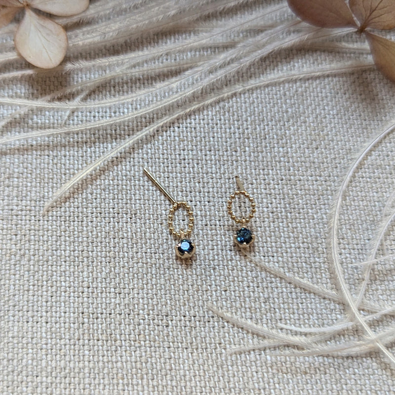 Gold earrings with dark gemstones on a textured beige surface with dried leaves.