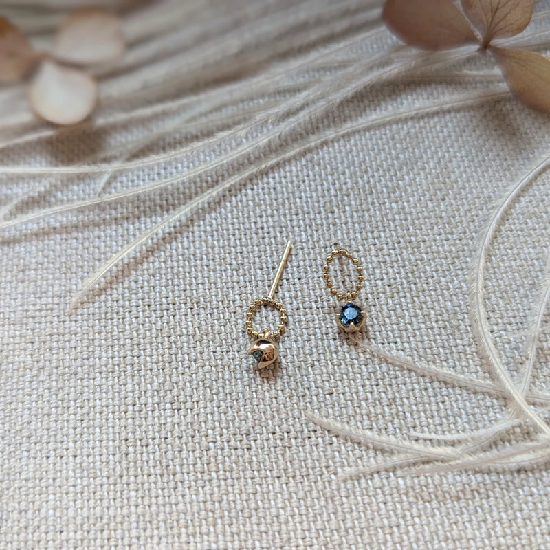 Pair of gold earrings with gemstones on a textured beige surface with dried leaves.