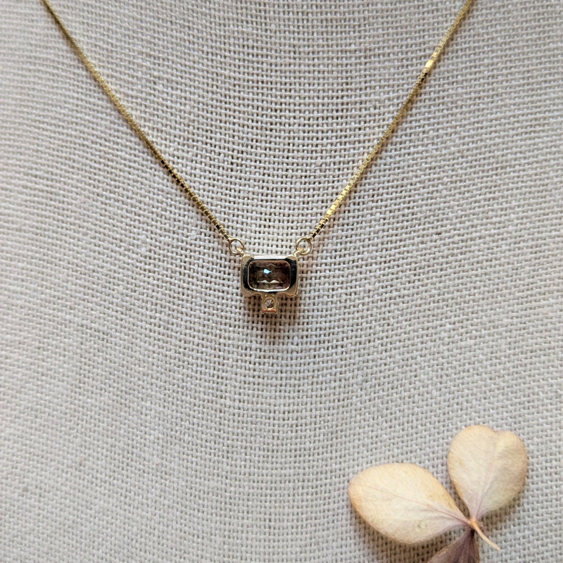 back of a gold necklace with a small pendant on a textured gray surface with a dried leaf.
