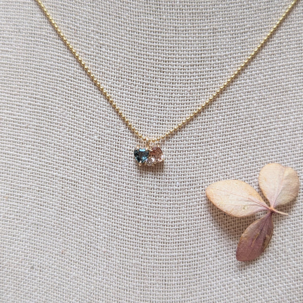 Gold necklace with a heart shaped blue gemstone and pink sapphire and diamond on a textured gray background with a dried leaf.