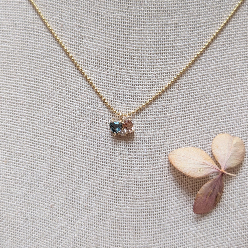 Gold necklace with a heart shaped blue gemstone and pink sapphire and diamond on a textured gray background with a dried leaf.