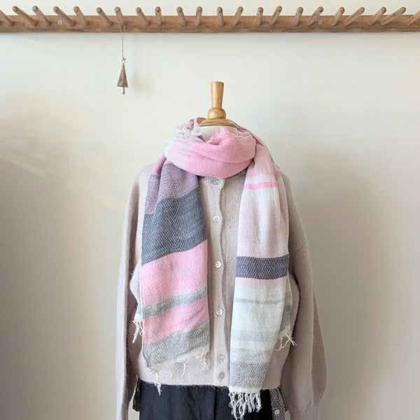 Scarf with pink and gray stripes draped over a beige cardigan on a wooden mannequin against a white wall.