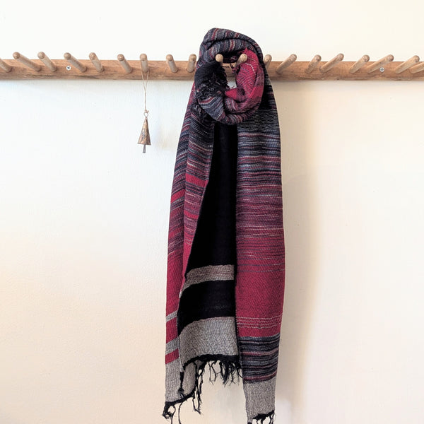 Striped scarf with red and black pattern draped over a wooden rack against a white wall.