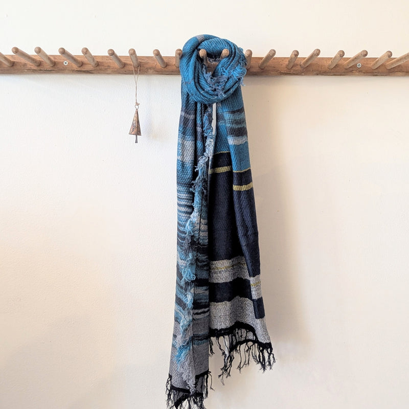 Blue and black striped scarf with tassels hanging on a wooden rack against a white wall.