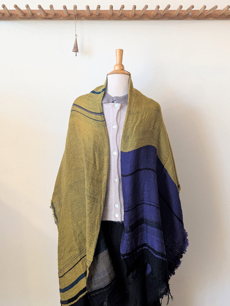 Mannequin wearing a green and blue striped shawl against a white wall.