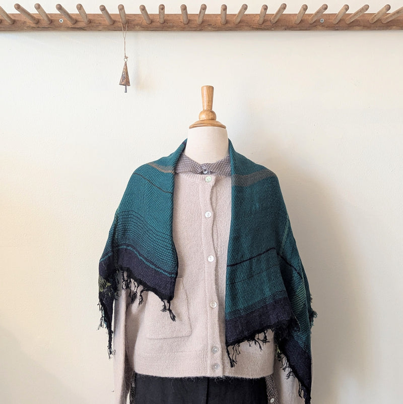 Mannequin wearing a beige sweater with a green and black patterned scarf draped over it, against a white wall.