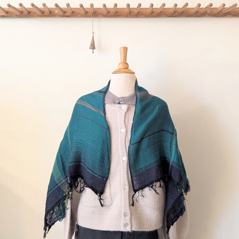 Mannequin wearing a teal and navy shawl over a beige shirt against a white background