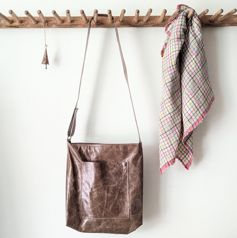 Brown leather bag hanging on a wooden rack with a plaid towel next to it.