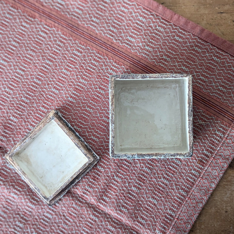 square ceramic box open with lid to the side on a textured red fabric surface