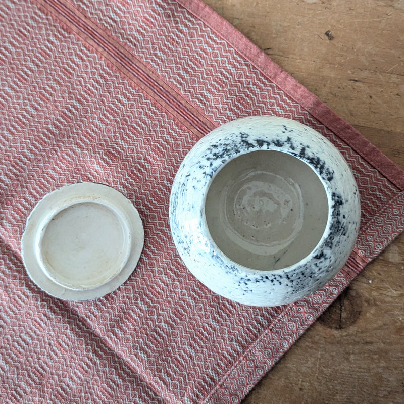 ceramic jar open with lid to the side on a textured red and white cloth with a wooden surface underneath.
