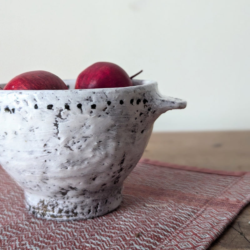 White ceramic bowl with black speckles containing red apples on a wooden surface with a pink woven mat.