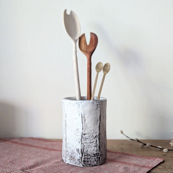 Set of wooden utensils in a textured ceramic holder on a wooden surface with a pink cloth.