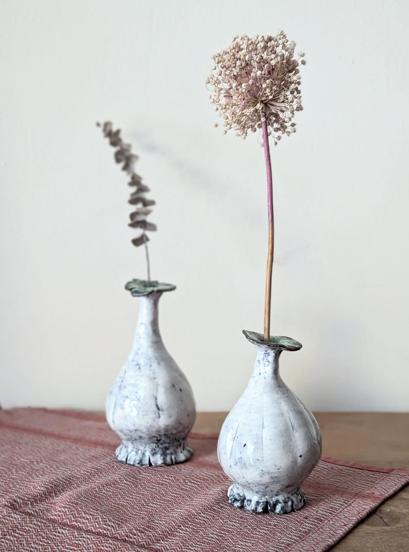 Two small white ceramic vases with dried plants on a textured surface.
