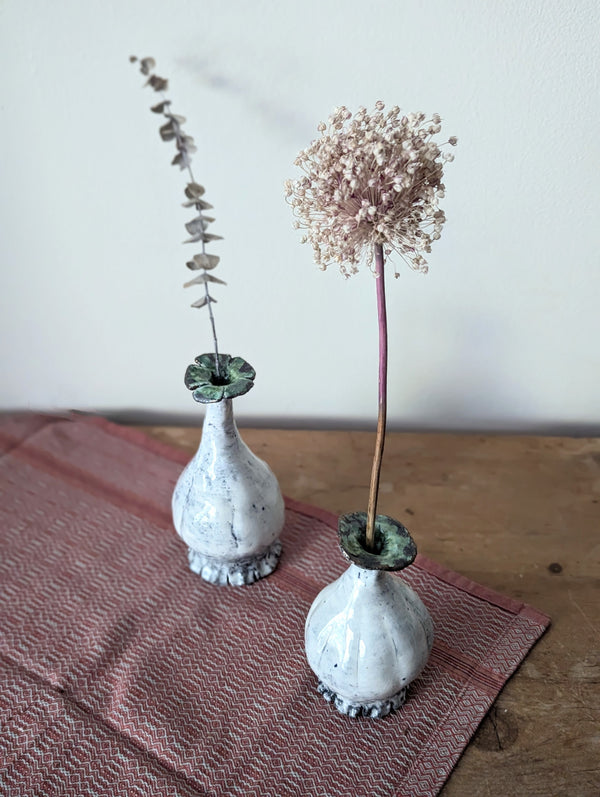 Two small ceramic vases with dried plants on a wooden surface with a pink cloth.