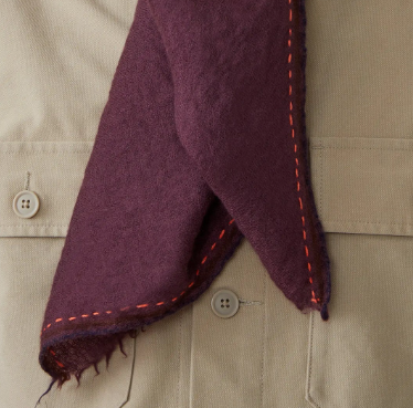 Close-up of a purple scarf with orange stitching on a beige garment. 